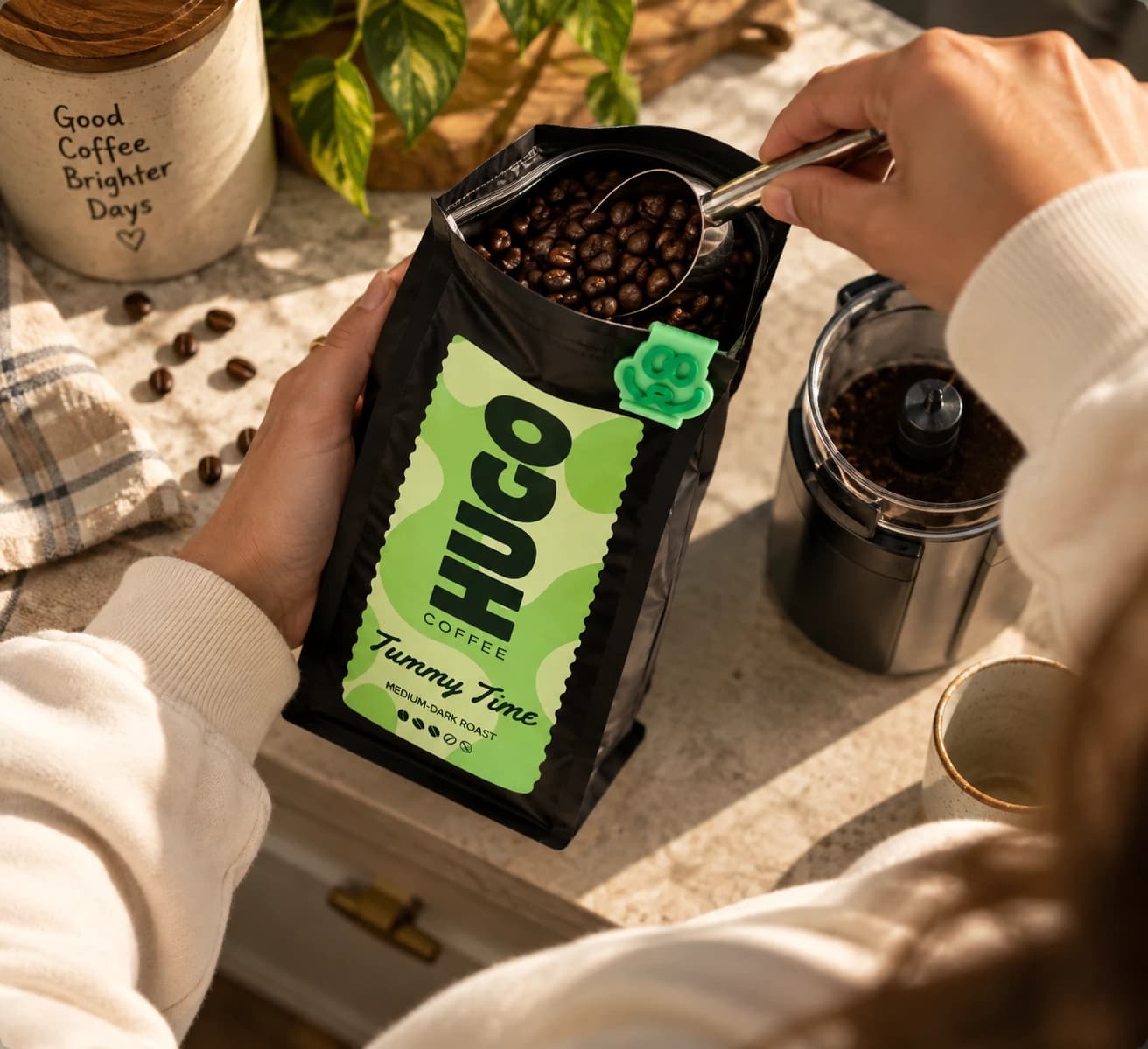 Whole beans scooped from a Tummy Time bag beside a grinder holding ground coffee